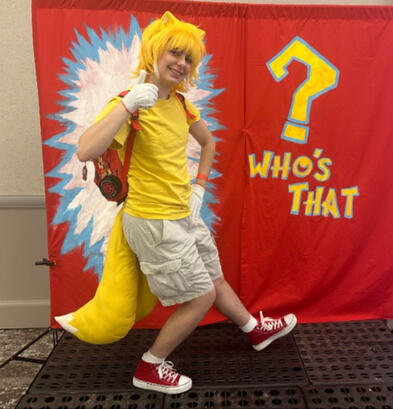 it's Tails Pine in Tails cosplay doing a thumbs up. standing in front of red backdrop with "who's that?" on the right side